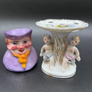 May include: Two antique ceramic figurines. One is a purple-colored head-shaped container with a smiling face and a yellow scarf. The other is a small, white, decorative stand with two cherubic figures and a floral design.