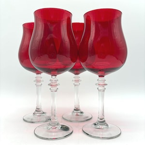 May include: Four red wine glasses with tulip-shaped bowls and clear stems and bases. The glasses are made of transparent red glass. The stems have a decorative design. The glasses are arranged in a group.