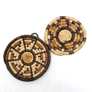May include: Two circular woven coasters or trivets featuring a concentric geometric pattern. One piece displays a dark brown, tan, and cream color palette, while the other features a lighter tan, beige, and dark brown design with a small loop.
