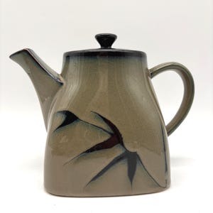 May include: A ceramic teapot with a rounded body, spout, and handle. The teapot is primarily a muted green color with a dark brown rim and lid. Dark brown and black floral design accents the body.