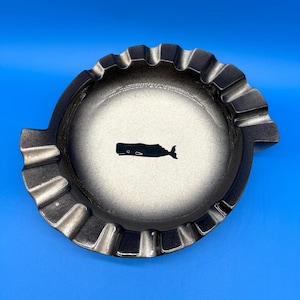 May include: A round ashtray with a whale silhouette in the center. The ashtray has a ruffled edge and a gradient color scheme, transitioning from black to white. The background is a solid blue.
