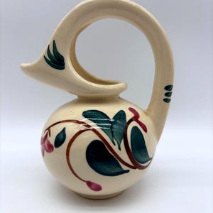 May include: Cream-colored ceramic pitcher with a distinctive arched handle. The pitcher is adorned with a floral design featuring green leaves and red flowers. The handle has a decorative element with green leaves.