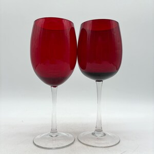 May include: Two wine glasses featuring deep red, translucent bowls and clear, slender stems that transition into circular, transparent bases. The glasses have a smooth, rounded shape and are positioned side-by-side against a plain background.