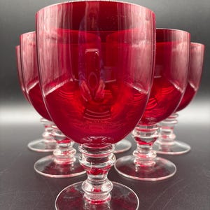 May include: A set of six ruby red wine glasses. The glasses have a clear, detailed stem and base, with a rounded bowl. The glasses are arranged in a group, with the light reflecting off the surfaces. The background is a dark gray.
