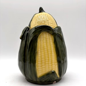 May include: A ceramic jar shaped like an ear of corn with a removable lid. The corn kernels are a pale yellow color, while the surrounding husks are a deep, glossy forest green. The piece features a textured surface with realistic details.