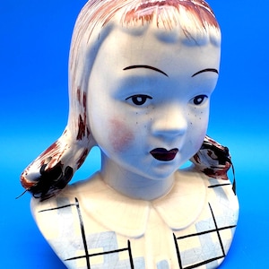 May include: A vintage ceramic girl head vase with a white and blue plaid design on the collar. The girl has brown hair, rosy cheeks, and dark lipstick. The vase is a unique decorative item.