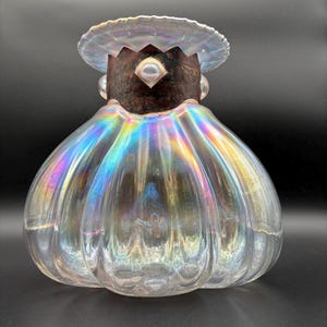 May include: A clear, iridescent glass decorative object with a bulbous body and a flared top. The glass has a ribbed texture and reflects a rainbow of colors. The top features a dark brown metal crown and a small, round pearl-like accent.