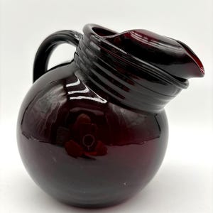 May include: A deep burgundy glass pitcher with a spherical body and a wide, curved spout. The neck features a series of horizontal ridges, and a smooth, dark handle is attached to the side. The glass has a glossy, reflective finish.