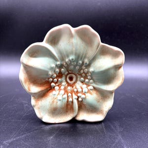 May include: A ceramic flower sculpture featuring five rounded petals with a pale mint green glaze. The center of the flower is detailed with raised, textured dots and a warm, rusty brown accent that radiates outward from the circular core.