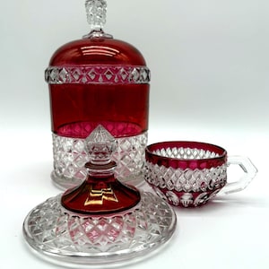 May include: A ruby red and clear cut glass lidded jar with a diamond pattern and a matching ruby red and clear cut glass teacup. The jar features a tall, pointed finial on its lid, while the teacup has a clear glass handle and a faceted base.