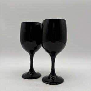 May include: Two matching wine glasses crafted from solid black glass. Each glass features a smooth, rounded bowl, a slender stem, and a circular base. The surface of the glass is opaque and reflective, showing a uniform dark finish throughout.