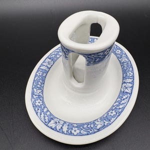May include: White ceramic ashtray with a blue floral design around the rim. The ashtray has a built-in rest for a cigarette. The design is a classic, vintage style.
