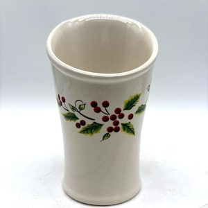 May include: A cream-colored ceramic tumbler featuring a hand-painted design of green holly leaves and clusters of small red berries encircling the upper portion of the vessel. The cup has a smooth, glossy finish and a slightly flared rim.