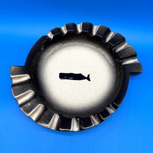 May include: A round, metallic ashtray featuring a gradient finish that transitions from a dark, charcoal-colored outer rim to a lighter, silver-toned center. A small, black silhouette of a whale is centered on the basin of the ashtray.