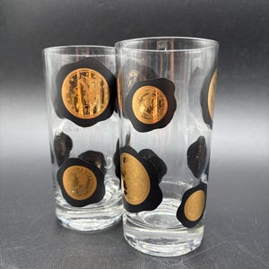 May include: Two clear glass tumblers decorated with black wax seal motifs featuring metallic gold coin designs. Each glass displays multiple circular black seals with intricate gold embossed imagery, creating a repeating pattern across the surface.