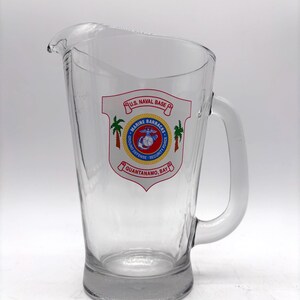 May include: Clear glass pitcher with a handle. The pitcher features a red, white, and blue emblem with text that reads "U.S. Naval Base Marine Barracks Guantanamo Bay". The emblem includes palm trees and a marine emblem.