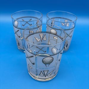 May include: Three clear drinking glasses with a golf theme. Each glass features a repeating pattern of golf balls, tees, clubs, and course markers in black and white. The glasses are set against a blue background.
