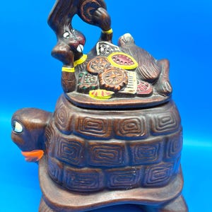 May include: A brown ceramic cookie jar in the shape of a turtle with a rabbit on top. The turtle has a textured shell and the rabbit is holding a tray of food. The jar has yellow and orange accents.