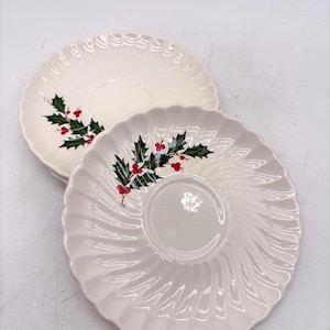 May include: Two off-white ceramic saucers with a scalloped, fluted edge design. Each saucer features a central circular indentation and a decorative illustration of green holly leaves with bright red berries positioned on the side of the plate.