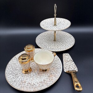 May include: A vintage tea set with a gold and white floral design. The set includes a two-tiered serving tray, a plate, a teacup, salt and pepper shakers, and a cake server. The items have gold trim and accents.