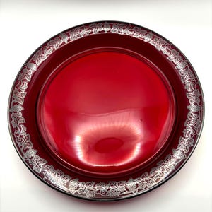 May include: A round, red glass serving tray with a silver floral design around the rim. The tray has a glossy finish and a black border. The decorative tray is suitable for serving food or as a decorative piece.