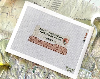 May include: A rectangular needlepoint canvas featuring a cross-stitch design of Rocky Mountain National Park, 1915. The design includes a brown mountain range, the park name in black text, and a small National Park Service arrowhead logo.