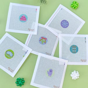 May include: A collection of square needlepoint canvases with various winter-themed designs, including a snowflake, a mitten, and a cupcake. The canvases are arranged on a light green surface with decorative bows and evergreen sprigs.