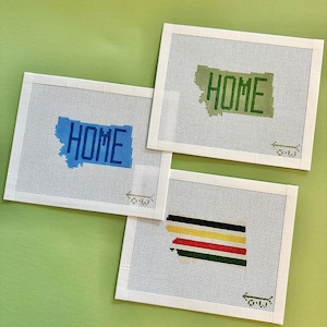 May include: Three needlepoint canvases with white borders and a grid pattern. Two canvases feature the state of Montana with the word "HOME" in blue and green. The third canvas has horizontal stripes in black, yellow, red, and green.