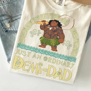 Puede incluir: Camiseta color crema con una ilustraci&oacute;n de dibujos animados de una figura polinesia sosteniendo un gran gancho. La camiseta tiene el texto "JUST AN ORDINARY DEMI-DAD" en amarillo. La camiseta est&aacute; doblada sobre una superficie azul claro con unos vaqueros y un sombrero.