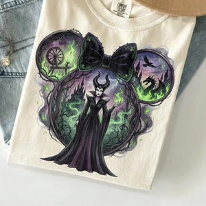 May include: Cream-colored t-shirt featuring a graphic of a Disney villain in a black dress, surrounded by a green and purple forest scene. The design is shaped like Mickey Mouse ears with a black bow. The shirt is folded on a light blue surface.