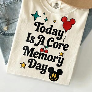 May include: Cream-colored t-shirt with the text "Today Is A Core Memory Day" in black, with a red Mickey Mouse balloon, stars, and cherries. The shirt is folded and placed on top of blue jeans and a tan hat.