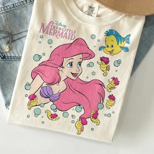 May include: Cream-colored t-shirt featuring a graphic of Ariel from Disney's The Little Mermaid. The design includes Ariel with pink hair, a purple seashell bra, and Flounder. The shirt also has seahorse and bubble illustrations.