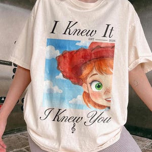 K&ouml;nnte beinhalten: Ein cremefarbenes Kurzarm-T-Shirt mit einem Grafikdruck einer Zeichentrickfigur mit roten Haaren und einem roten Hut vor einem blauen Himmel mit Wolken. Der Text "I Knew It" steht &uuml;ber der Grafik und "I Knew You" ist darunter gedruckt.
