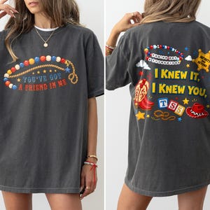 Pu&ograve; includere: Una t-shirt grigio antracite a maniche corte con una grafica frontale di una collana di perline colorate e il testo "YOU'VE GOT A FRIEND IN ME". Il retro mostra una grafica pi&ugrave; grande con stivali da cowboy, un cappello, blocchi e il testo "I KNEW IT, I KNEW YOU".