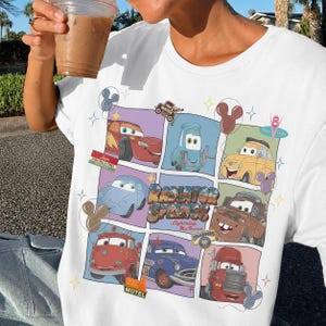 May include: A white t-shirt featuring a grid of colorful illustrations of characters from the movie Cars, including Lightning McQueen, Mater, and Luigi. The center text reads Radiator Springs Lightning McQueen, surrounded by small star and balloon icons.