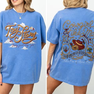 May include: A blue short-sleeve t-shirt featuring a front graphic with the text "it's a Toy Story" in rope-style lettering over clouds. The back graphic displays "Cowgirl like Me" and "Takes One to Know One" with a hat, rope, and desert imagery.