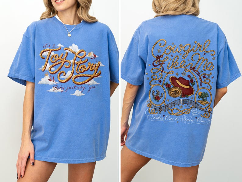May include: A blue short-sleeve t-shirt featuring a front graphic with the text "it's a Toy Story" in rope-style lettering over clouds. The back graphic displays "Cowgirl like Me" and "Takes One to Know One" with a hat, rope, and desert imagery.