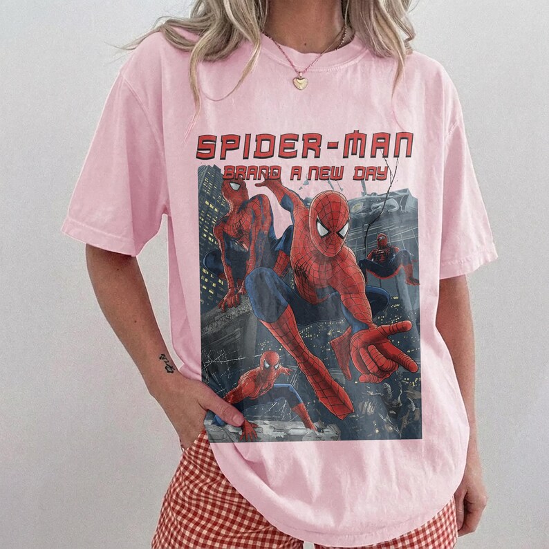 May include: A light pink short-sleeve t-shirt featuring a graphic print of Spider-Man in various action poses against a dark city background. The text "SPIDER-MAN BRAND A NEW DAY" is printed in red block letters above the central character illustration.