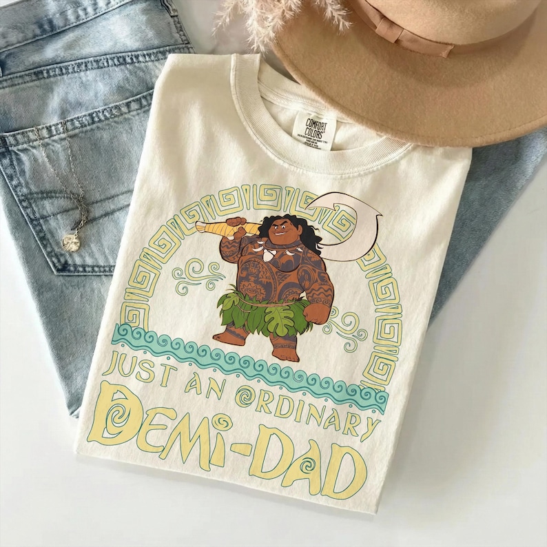 Puede incluir: Camiseta color crema con una ilustraci&oacute;n de dibujos animados de una figura polinesia sosteniendo un gran gancho. La camiseta tiene el texto "JUST AN ORDINARY DEMI-DAD" en amarillo. La camiseta est&aacute; doblada sobre una superficie azul claro con unos vaqueros y un sombrero.