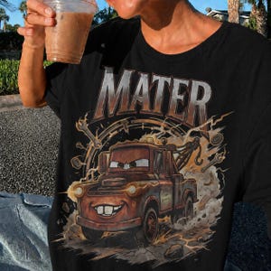 May include: Black t-shirt featuring a graphic of Mater from the movie Cars. The design includes the word "MATER" in large, metallic letters above an illustration of the character. The image has a distressed, vintage look with lightning bolts and tools.