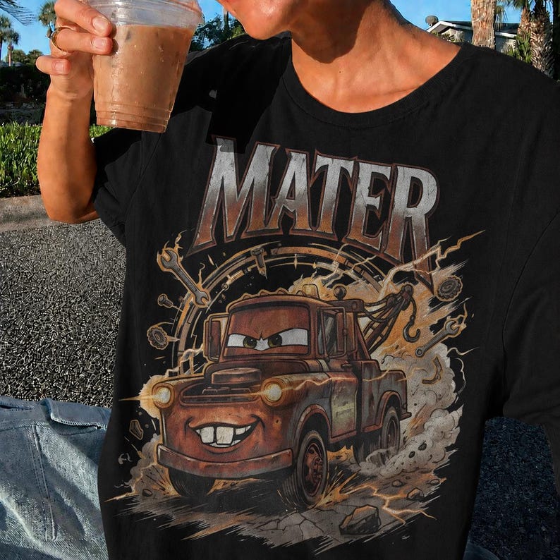 May include: Black t-shirt featuring a graphic of Mater from the movie Cars. The design includes the word "MATER" in large, metallic letters above an illustration of the character. The image has a distressed, vintage look with lightning bolts and tools.