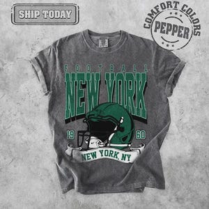 May include: Gray t-shirt with a distressed look, featuring the words "FOOTBALL NEW YORK" in green block letters. A green football helmet is centered below the text, with the years "19" and "60" on either side. The words "NEW YORK, NY" are on a banner.