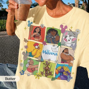 Op de afbeelding: Boterkleurig T-shirt met een rasterontwerp van Moana-personages. Het ontwerp bevat illustraties van Moana, Maui, Pua, Hei Hei en anderen, met het Disney Moana-logo. Het shirt heeft een klassieke ronde hals en korte mouwen.