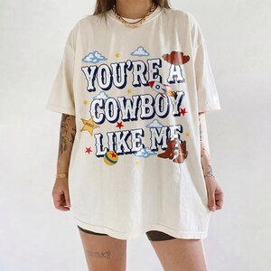 Puede incluir: Una camiseta de manga corta color hueso con el texto "YOU'RE A COWBOY LIKE ME" en letras azules. El gr&aacute;fico incluye un cohete, un sombrero de vaquero, un par de botas marrones, una estrella amarilla, una pelota colorida y nubes y estrellas.