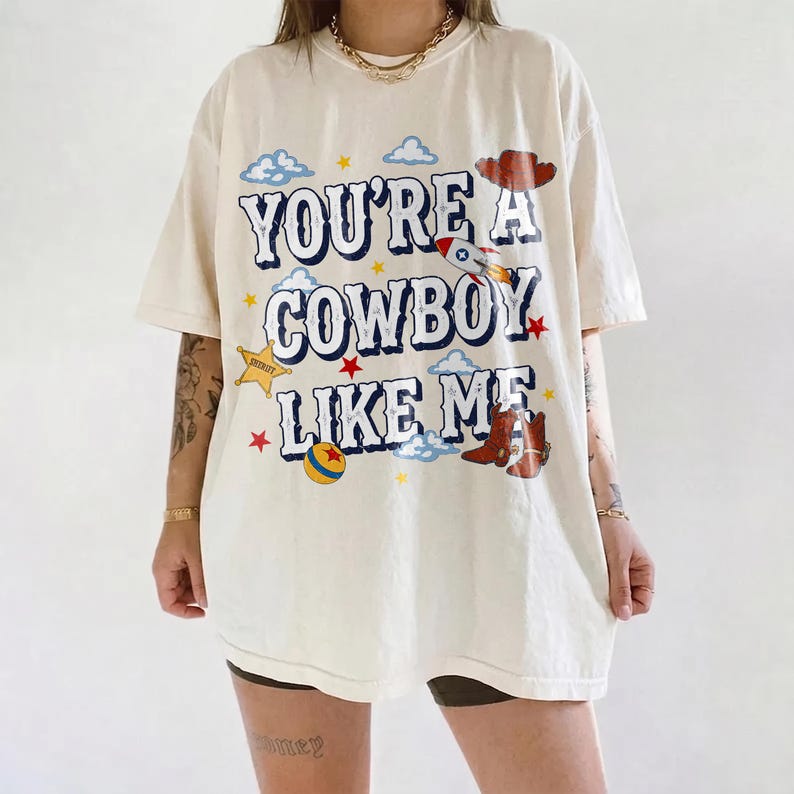 Puede incluir: Una camiseta de manga corta color hueso con el texto "YOU'RE A COWBOY LIKE ME" en letras azules. El gr&aacute;fico incluye un cohete, un sombrero de vaquero, un par de botas marrones, una estrella amarilla, una pelota colorida y nubes y estrellas.