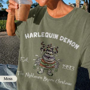 May include: Moss green t-shirt with the text "HARLEQUIN DEMON" and a cartoon mummy-like character. The shirt also features the text "Est. 1993" and "The Nightmare Before Christmas".