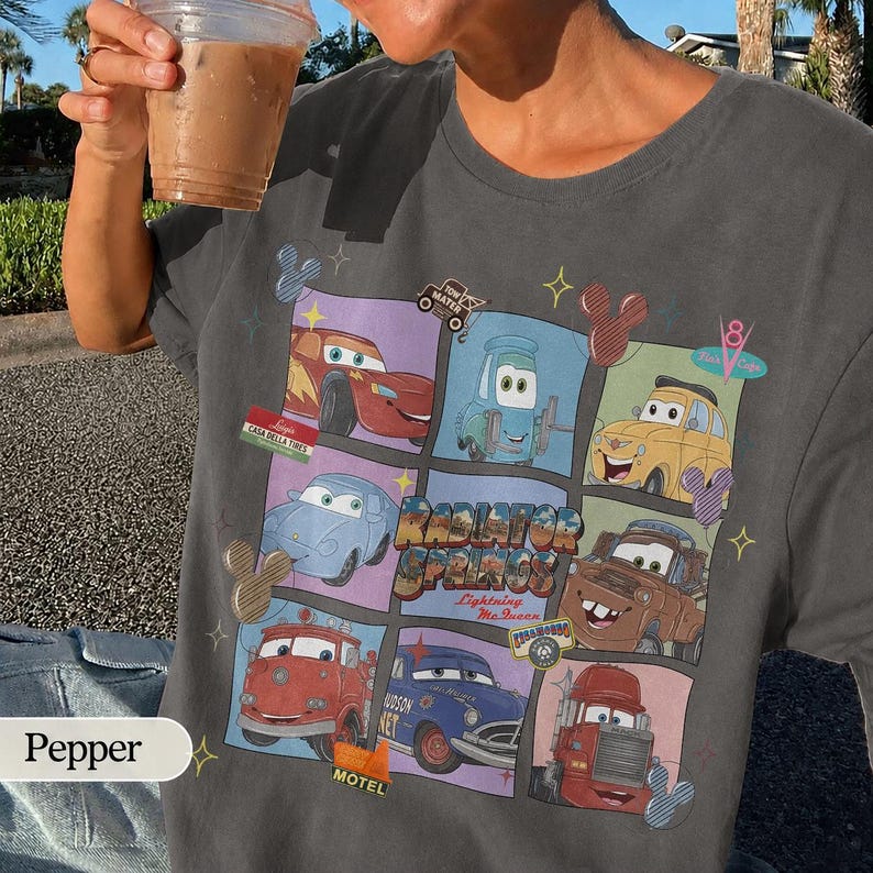 May include: A charcoal gray t-shirt featuring a grid graphic of various characters from the movie Cars, including Lightning McQueen, Mater, and Luigi. The design includes the text Radiator Springs and Lightning McQueen, accented with small star and mouse ear icons.