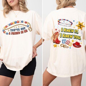 Pu&ograve; includere: Una t-shirt color crema con una grafica frontale di una collana di perline colorate e il testo "YOU'VE GOT A FRIEND IN ME". La grafica sul retro mostra una collana di perline, una stella da sceriffo, stivali da cowboy, un cappello e il testo "I KNEW IT, I KNEW YOU".