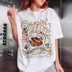 May include: A white short-sleeve t-shirt featuring a graphic design with the text Cowgirl Like Me, Eyes full of Stars, and Takes One to Know One. The illustration includes a brown cowboy hat, a horseshoe, a cactus, roses, and rope motifs.