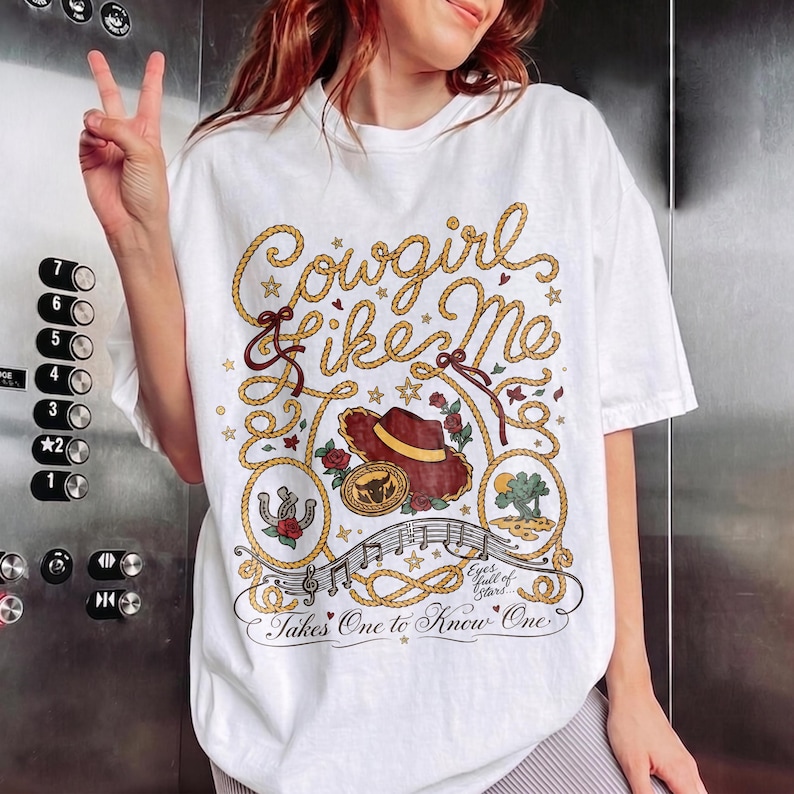 May include: A white short-sleeve t-shirt featuring a graphic design with the text Cowgirl Like Me, Eyes full of Stars, and Takes One to Know One. The illustration includes a brown cowboy hat, a horseshoe, a cactus, roses, and rope motifs.